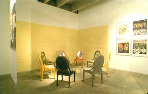 Panorama of an art installation: seven rounded-back chairs circle at center, a hanging banner to the left, a golden wall with text in the background and four framed artworks to the right.