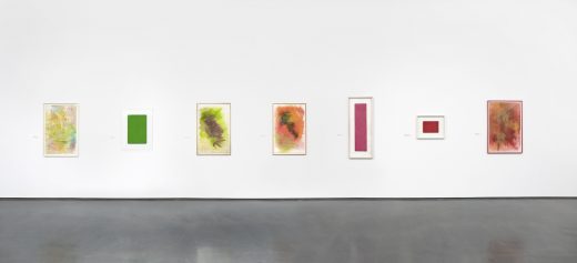 Seven colorful, abstract paintings of different sizes hang in a row on a white gallery wall.