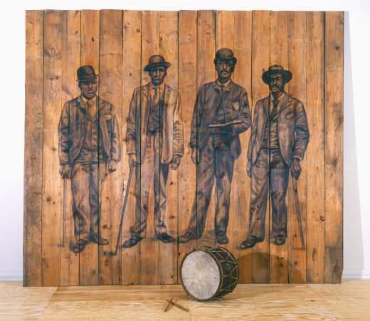 Charcoal drawing of four besuited Black men on a wooden panel with an old drum and drumsticks. 