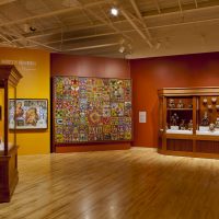 Alt: A museum gallery showcasing Mesoamerican pottery, paintings and tapestries against a red wall.