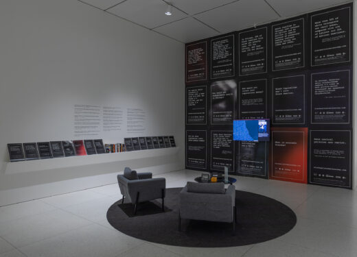Gallery installation showing a TV monitor on a wall covered with posters and a pair of chairs