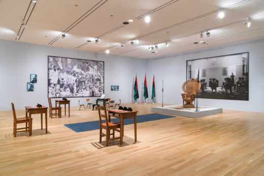 Gallery installation consisting of school desks, a woven throne, flags, and large scale photographs.