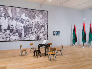 Gallery installation with a small piano, three flags, and a large scale photograph.