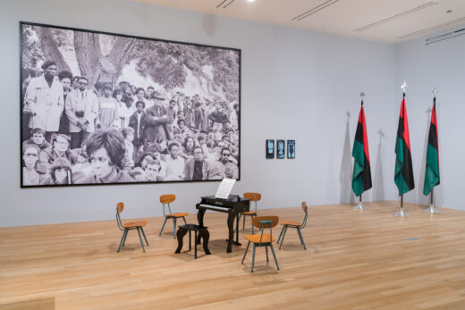 Gallery installation with a small piano, three flags, and a large scale photograph.