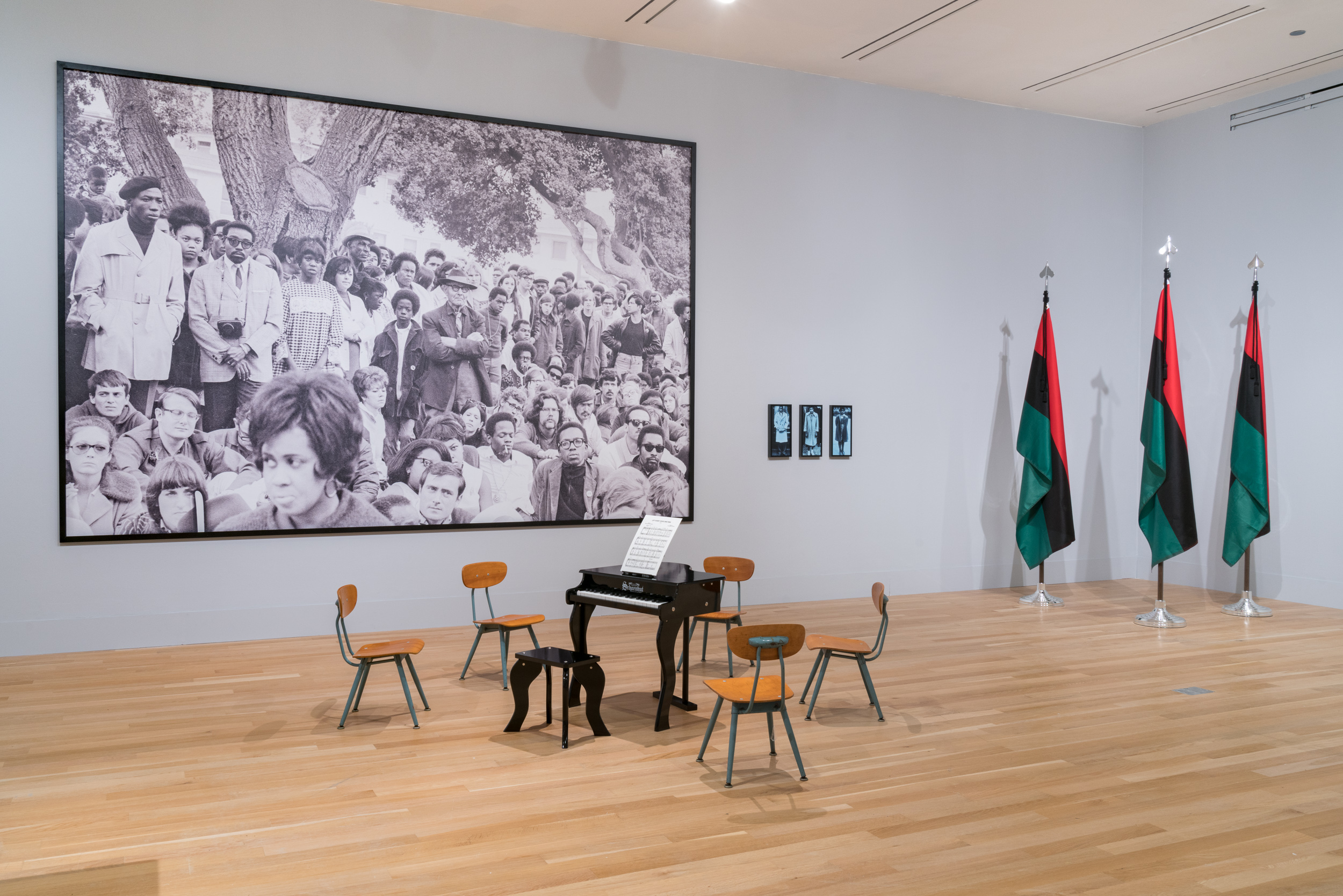 Gallery installation with a small piano, three flags, and a large scale photograph.