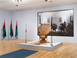 Gallery installation featuring a woven throne, three flags, and a large scale photograph of a classroom.