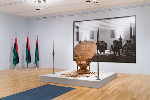 Gallery installation featuring a woven throne, three flags, and a large scale photograph of a classroom.