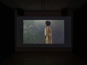 Gallery black box theatre showing a film of a woman standing in a yellow kimono.
