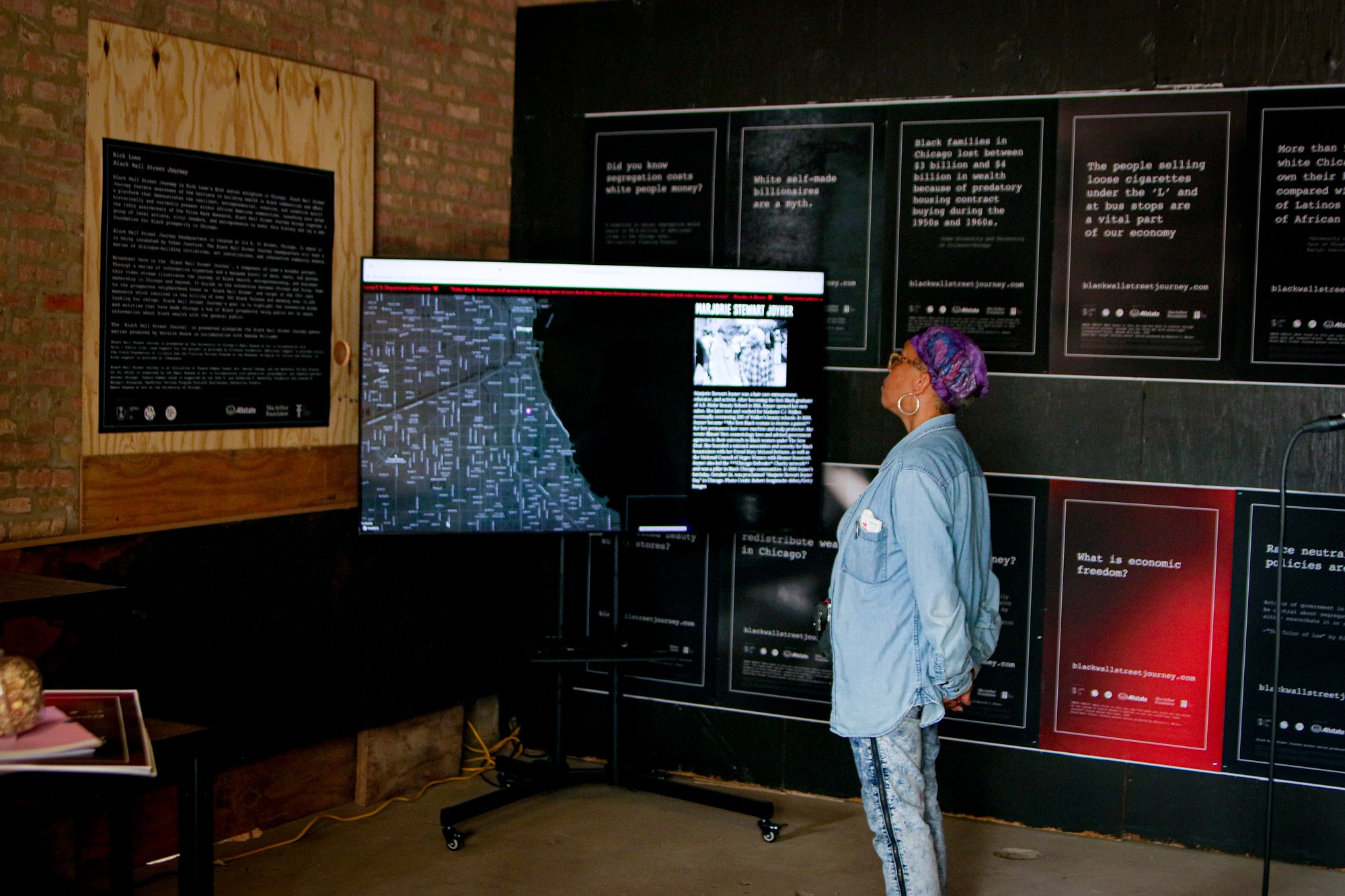 A woman in denim watches a TV monitor mounted in from of posters in a gallery exhibition.