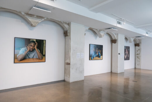 Gallery installation of three large color photographs of individual students seated at desks.