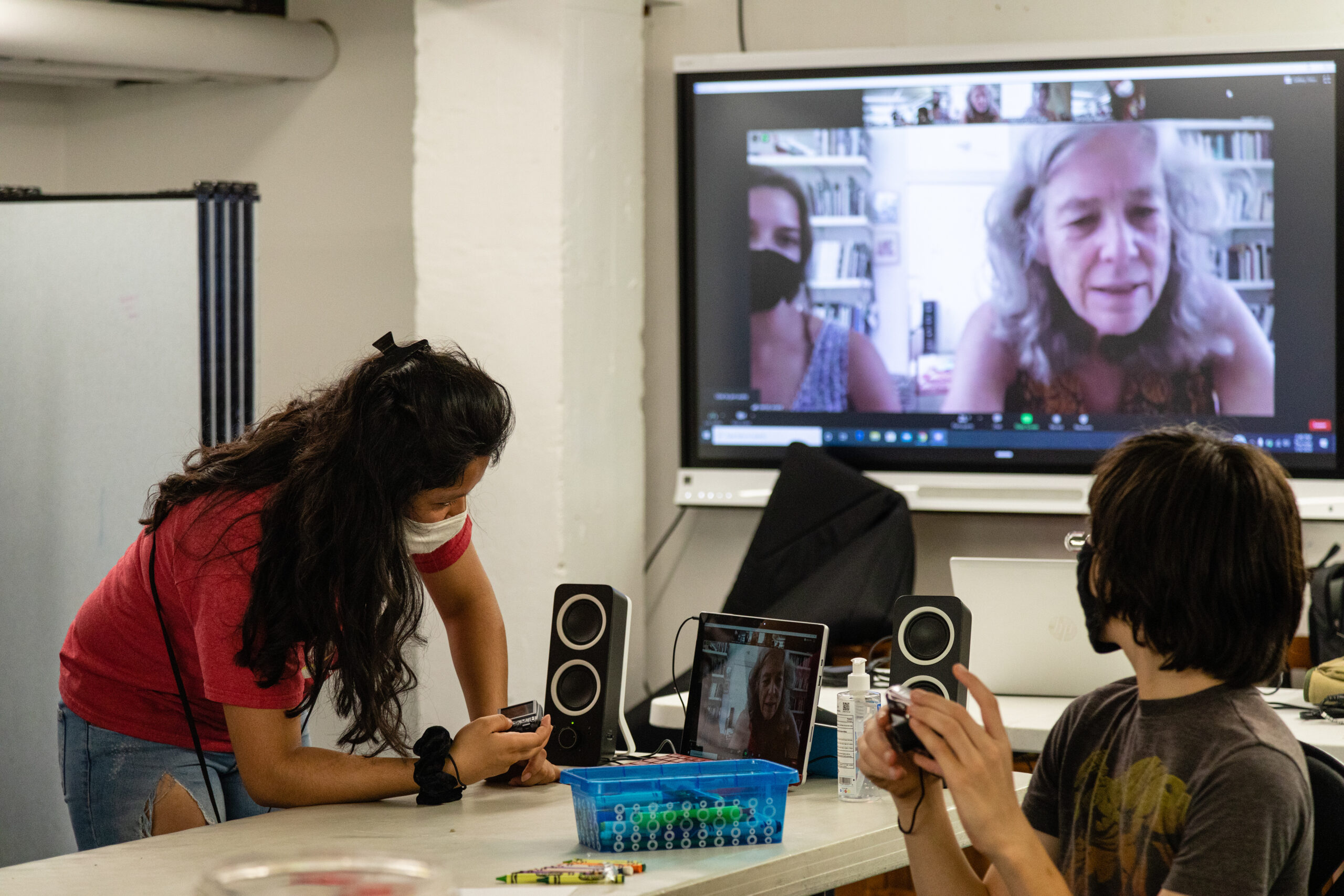 Two youth, wearing masks, work with hand-held cameras while other participate remotely via a wall-mounted monitor