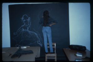 The artist stands on a chair, drawing in chalk on a large piece of black paper