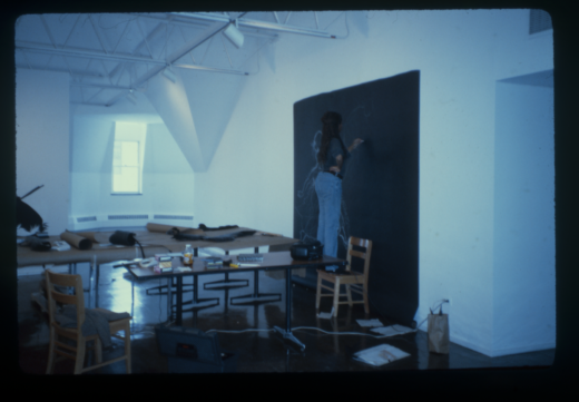 The artist stands on a chair to make a large chalk drawing on black paper