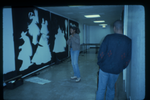 Two people in a gallery hallway, one leans against the wall and one stands laughing