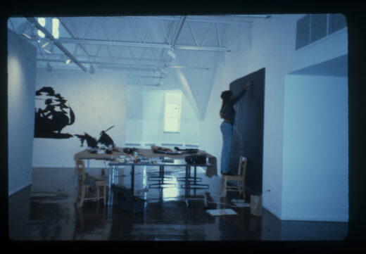 The artist stands on a chair, drawing on a large black piece of paper in white chalk, with an artwork in the same style installed on the wall behind.