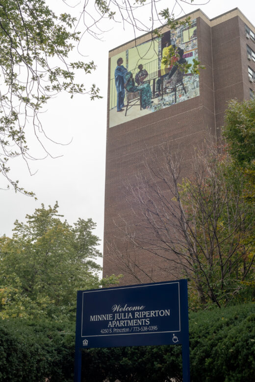 A tall brick building with a vinyl mural depicting a domestic scene of five people gathered around a kitchen table