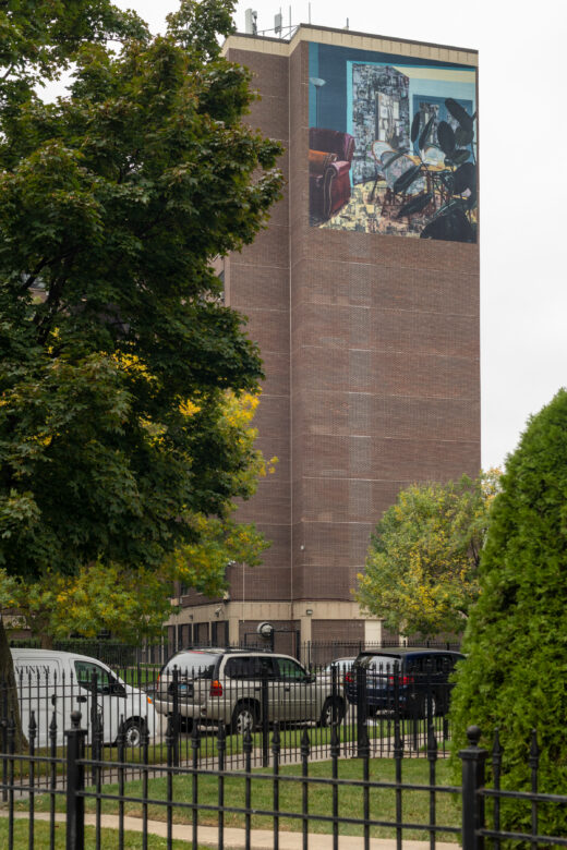 A tall brick building with a vinyl mural depicting a domestic scene of an empty living room with a rubber plant