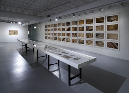 An installation with a grid of photographs depicting aerial vantages of the desert with white vitrine tables in the foreground