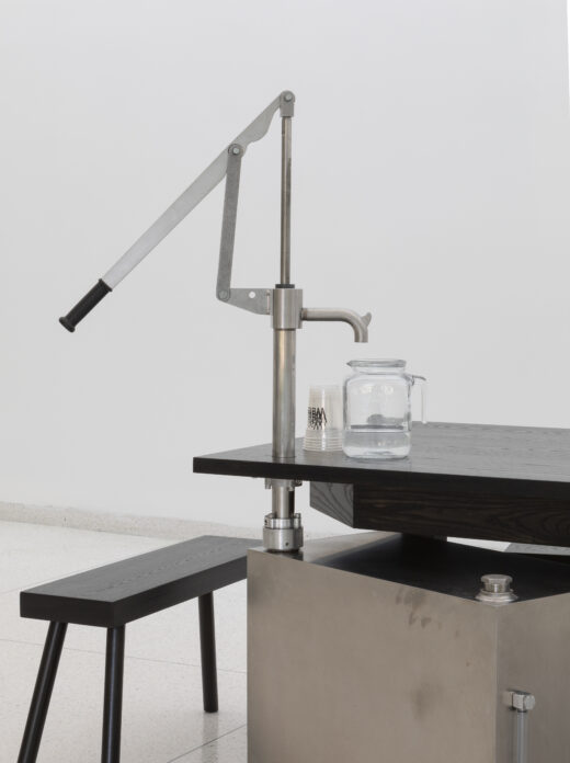 A metal hand pump fixed to the corner of a table brings water to a glass jar